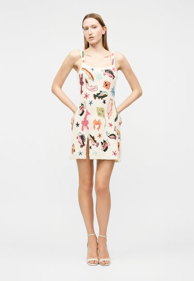 Γυναίκα που φοράει λευκό μίνι φόρεμα χωρίς μανίκια με πολύχρωμα κεντήματα θαλάσσιων πλασμάτων και αστεριών, στέκεται με τα χέρια στη μέση μπροστά σε λευκό φόντο.