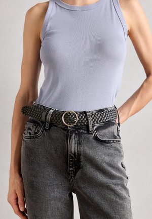 Ceinture noire tissée avec une boucle ronde dorée, portée sur un jean gris taille haute et un top sans manches lilas, mettant en avant la texture et le design.