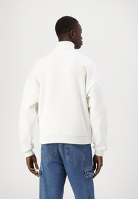 Hvit oversized sweatshirt med høy krage, ribbestrikkede mansjetter og kant; kombinert med blå denimbukser med lommer og sømdetaljer.
