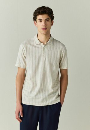 Młody mężczyzna z krótkimi kręconymi włosami, ubrany w beżową, pasiastą koszulkę polo z krótkim rękawem i ciemne spodnie, stojący na jednolitym jasnym tle.