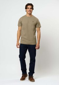 Beige puuvillane T-särk ümmarguse kaeluse ja lühikeste varrukatega, millel on rindade kohal väike must logo, kombineeritud tumehallide teksade ja pruunide saapadega.