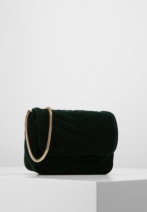 Czarna pikowana aksamitna torba na ramię ze złotym łańcuszkiem, spoczywająca na białej powierzchni na tle jasnego, jednolitego tła.