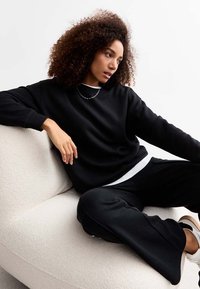 Sudadera de algodón negra con un corte relajado, cuello redondo y puños acanalados, combinada con pantalones de pierna ancha negros sobre un fondo claro texturizado.