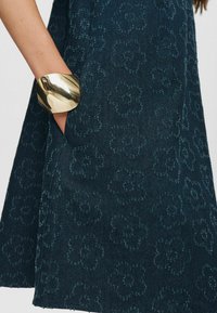 Mörkblå klänning med blommönster och texturerad yta, som har en sidoficka och ett stort guldmetallarmband på handleden.