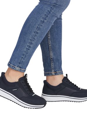 Person iført mørkeblå træningssko og blå skinny jeans, benene krydset ved anklerne mod en hvid baggrund.