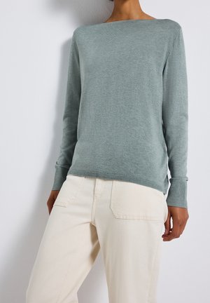 Persona che indossa un maglione a maniche lunghe azzurro chiaro e pantaloni color crema, in piedi davanti a uno sfondo bianco semplice.