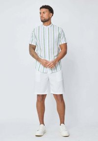 Chemise à manches courtes, à boutons, avec des rayures verticales vertes et blanches, portée avec un short blanc doté de deux poches avant et des baskets blanches.