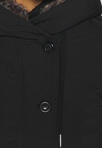 Svart pufferjacka med en huva klädd i päls, två svarta knappar och dragsko detaljer vid halsen. Slät yttermaterial med en matt finish.