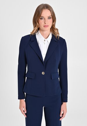 Blazer blu navy con un taglio aderente, due tasche frontali, un singolo bottone dorato e rever a punte, abbinato a una camicia bianca con colletto.