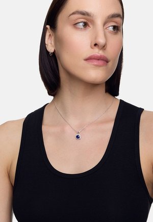 Srebrna naszyjnik z kwadratowym niebieskim zawieszeniem, osadzonym w okrągłej ramie, noszony z czarnym topem na ramiączkach. Modelka ma krótkie brązowe włosy.