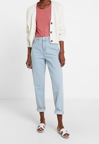 Lyserød tanktop under en cremet, ribbet cardigan med knapper, parret med lys blå denimjeans med opkrøllede kanter og hvide slidesandaler.