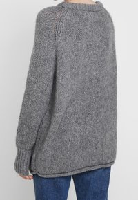 Persone che indossa un maglione grigio chunky a maglia larga con maniche lunghe e polsini a coste, abbinato a jeans di denim blu, visto da dietro.