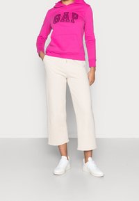 Felpa con cappuccio fucsia con logo "GAP", abbinata a pantaloni della tuta beige a vita alta e sneakers bianche. Outfit casual e comodo.