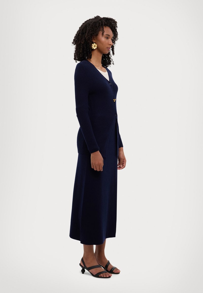 Cardigan a coste navy con chiusura a bottoni, dotato di scollatura a V e orlo leggermente svasato, indossato sopra un top chiaro e abbinato a sandali neri.
