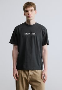 Pelēka kokvilnas T-krekls ar apaļu kakla izgriezumu, priekšpusē uzkrāsots balts logo "CALVIN KLEIN NEW EST. 1985 YORK".