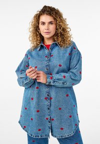 Sinine denim-pusa pikkade varrukatega, esitleb punaste huuleprindiga aktsente, nööbiga sulgemine ja ümar äär, kantud punase t-särgi peal.