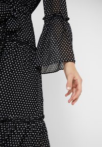 Main à la peau claire portant une robe noire à pois blancs avec de longues manches amples et transparentes sur fond blanc.