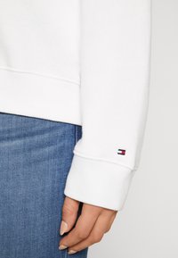 Sudadera blanca con puños acanalados y un pequeño parche de logo multicolor en la manga, combinada con jeans de denim azul.