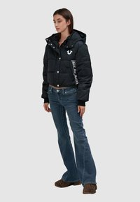 Czarna skrócona kurtka puchowa z kapturem, z zatrzaskami i białym logo "True Religion". Noszona z dżinsami w linii dzwon i butami w zwierzęcy wzór.