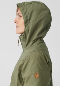 Olivgrön huvjacka i slätt tyg. Har dragkedja framtill och en brun logotyp på vänster ärm.