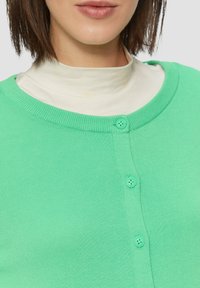 Minzgrüner Cardigan mit rundem Ausschnitt, gerippten Kanten und drei Knopfverschlüssen auf der Vorderseite, über einem cremefarbenen Oberteil getragen.