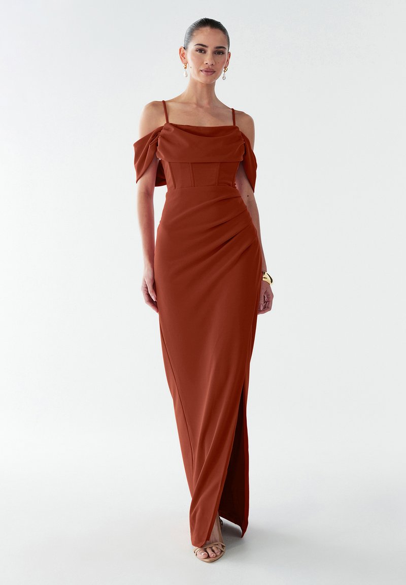 Nais wearing rust-oranži off-shoulder kleit õhukeste rihmadega, külgsilmaga, kuldse käevõruga, pärlidega kõrvarõngastega ja beežide kontsadega, seismas valgel taustal.
