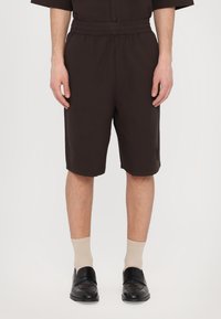 Brown, knee-length shorts with an elastic waistband, featuring a textured fabric and a side pocket, paired with beige socks and black shoes.