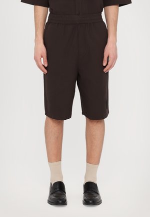 Brown, knee-length shorts with an elastic waistband, featuring a textured fabric and a side pocket, paired with beige socks and black shoes.