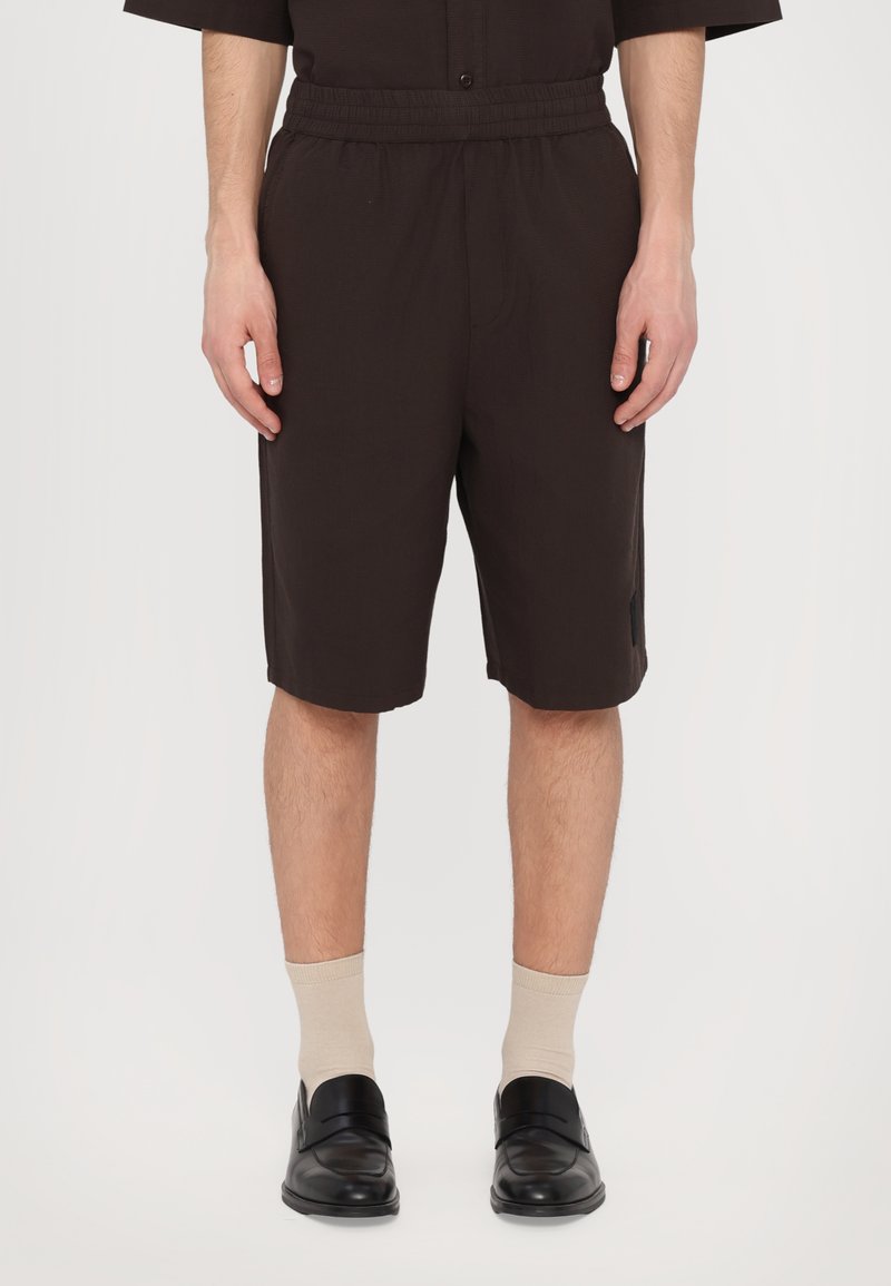Brown, knee-length shorts with an elastic waistband, featuring a textured fabric and a side pocket, paired with beige socks and black shoes.