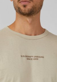 Beige katoenen t-shirt met een ronde halslijn, voorzien van bruine bedrukte tekst "S.OLIVER® UNBOUND SINCE 1969" op de borst.