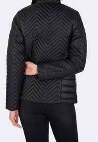 Veste rembourrée noire avec un design matelassé en chevrons, des zips et un col montant. Le dos présente une silhouette ajustée et des poches latérales.