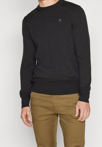 Pull noir en maille, col rond, poignets et ourlet côtelés. Petit détail logo sur la poitrine, associé à un pantalon beige.