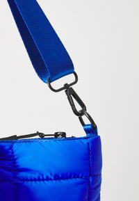 Sac matelassé bleu avec une finition brillante. Il dispose d'une large sangle en tissu et d'un clip en métal noir, avec un haut zippé et des détails de couture.