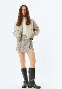 Beige bomber jacketin päällä vaalean crème neuleen, yhdistettynä kuvioituun käärmeennahkaskorttiin ja polvipitkiin mustiin saappaisiin, joissa on soljet.