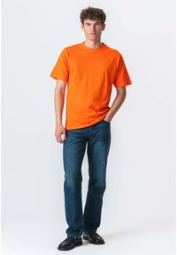 En ung mand står iført en lys orange t-shirt, blå jeans og sorte sko foran en ensfarvet hvid baggrund.