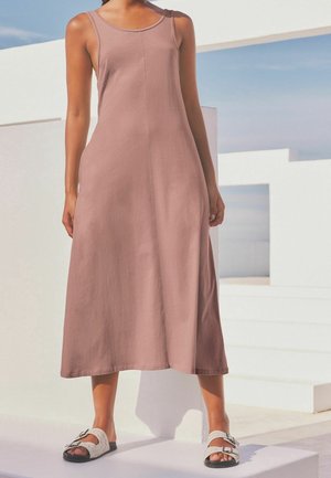 Mujer de pie al aire libre, vistiendo un vestido midi sin mangas color rosa polvoriento y sandalias blancas con hebillas, contra un fondo arquitectónico blanco.