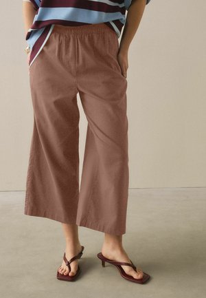 Persoon draagt bruine broek met wijde pijpen en korte lengte en bordeauxkleurige sandalen met hakken, staand op een gladde beige vloer tegen een effen muur.