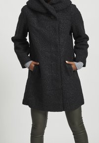 Svart texturerad kappa med hög krage, knappslutning och sidofickor; tillsammans med grå stickade manschetter och åtsittande gröna läderbyxor.