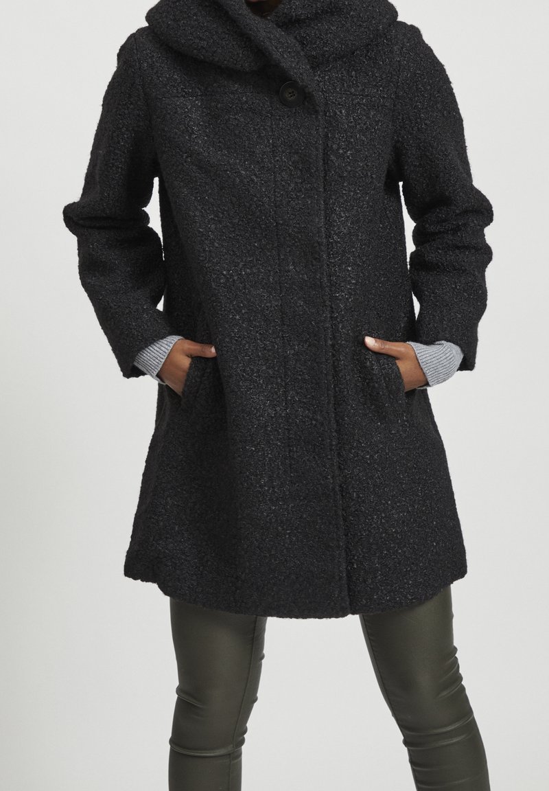 Svart texturerad kappa med hög krage, knappslutning och sidofickor; tillsammans med grå stickade manschetter och åtsittande gröna läderbyxor.