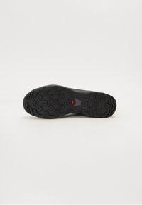 Czarna podeszwa sportowego buta z teksturowanym wzorem, posiadająca okrągłe uchwyty i wyraźny czerwony trójkątny akcent. Materiał gumowy zapewniający przyczepność.