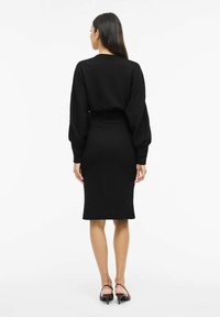 Musta neulemekko, jossa on vartalonmyötäinen siluetti, pitkät pussihihat ja resorivyötärö. Sileä tekstuuri, polvipituinen, yhdistetty korkeakorkoisten kenkien kanssa.