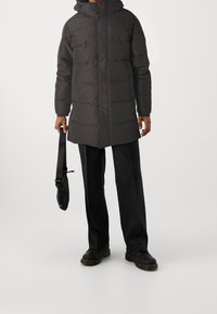 Lunga giacca a vento nera con zip frontale, design trapuntato e cappuccio. Il modello tiene in mano una borsa nera e indossa pantaloni e scarpe neri.