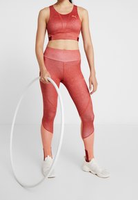 Brassière de sport rouge avec un design sportif et des accents en maille, associée à un legging dégradé assorti. Le modèle tient un cerceau blanc. Chaussures de sport blanches.