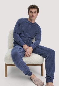 Navyblaues Loungewear-Set aus weichem Material, mit langen Ärmeln und elastischen Bündchen, kombiniert mit leichten Hausschuhen. Modell sitzt auf einem texturierten Stuhl.