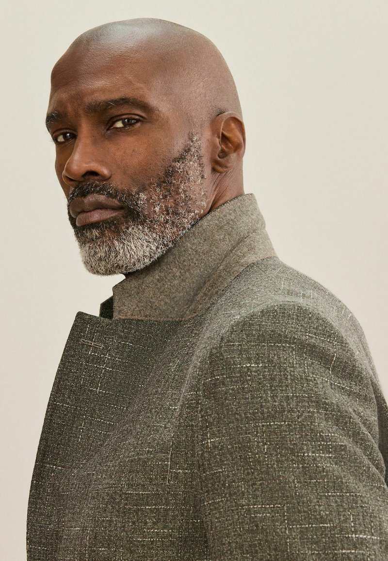 Homme mûr et chauve avec une barbe grise portant un manteau gris texturé, regardant par-dessus son épaule sur un fond uni.