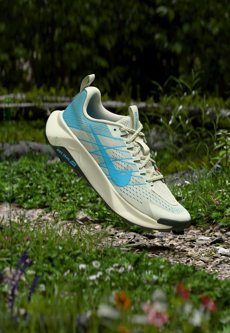 Lys grønn og blå Nike terrengløpesko svever over en gresskledd, steinete sti med villblomster og skog i bakgrunnen.