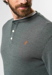 Pilkas langeliais ilgomis rankovėmis marškinis su mygtukais henley apykakle. Su mažuoju oranžiniu išsiuvinėtu logotipu ant krūtinės. Tekstūruota medžiaga.