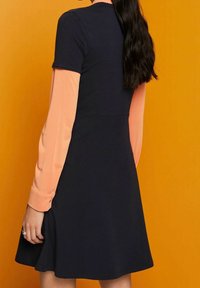 Granatowa sukienka krótko rękawowa z rozkloszowanym dołem, nałożona na różową bluzkę z długim rękawem; ma okrągły dekolt i miękką teksturę materiału.