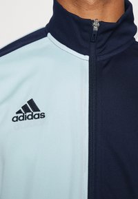 Adidas träningsjacka i ljusblått och marinblått med hög krage, dragkedja och svart logotyp på vänster bröst. Slät textur och sportig design.