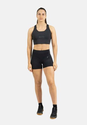 Mujer de pie mirando hacia adelante, vistiendo un sujetador deportivo negro, pantalones cortos negros de talle alto, calcetines tobilleros negros y zapatillas deportivas negras.
